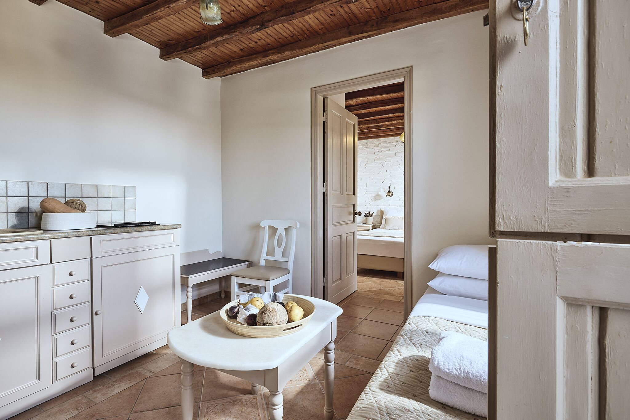 A cozy, well-lit room with a wooden ceiling, a white tiled backsplash, a dining table with a basket of fruits, a white sofa, and a bed with white linens.