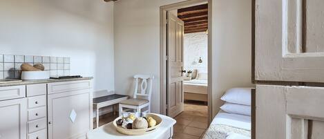 Una habitación acogedora y bien iluminada con techo de madera, un rebozado blanco, una mesa de comedor con una cesta de frutas, un sofá blanco y una cama con ropa de cama blanca.