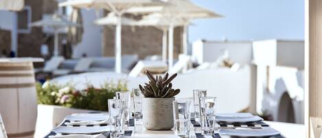 A table set for dining with a potted plant, glasses, and cutlery, overlooking a pool area with lounge chairs and umbrellas.