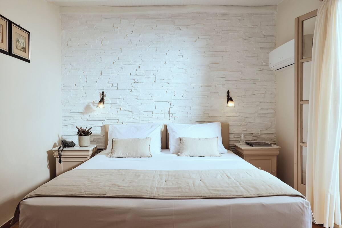 A hotel room with a white brick wall, a bed with white linens, two bedside lamps, a nightstand with a phone and a plant, and a window with curtains.