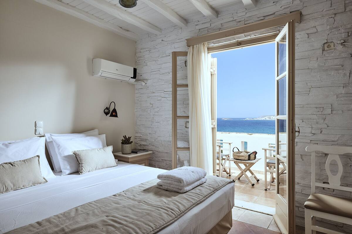 A bedroom with a bed, a nightstand, a lamp, and a view of the beach through an open door.