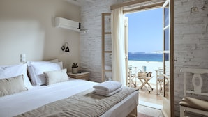 A bedroom with a bed, a nightstand, a lamp, and a view of the beach through an open door. - Naxos Magic Village Hotel (Naxos)
