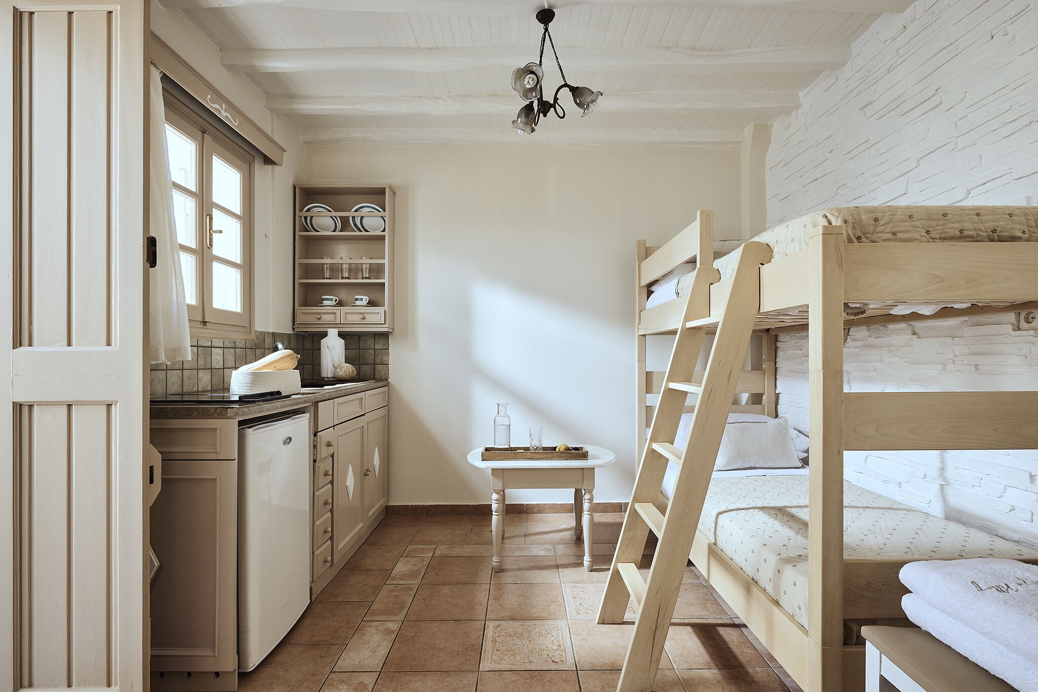 A compact kitchen with a wooden table, a built-in shelf with dishes, and a bunk bed with a ladder.