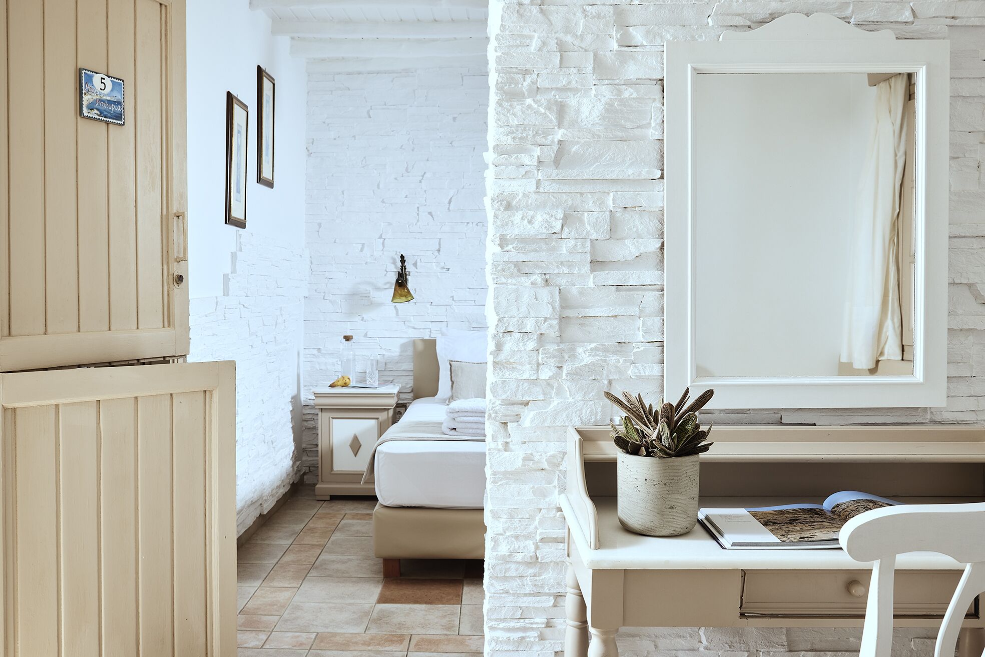Une pièce avec un mur en briques blanches, un lit, une table de chevet avec une lampe, une plante en pot et un miroir.