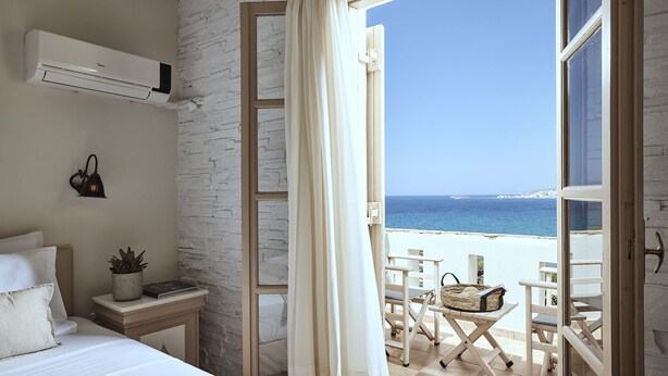 A bedroom with a view of the beach, a bed, a bedside table with a plant, and a window with curtains. - Naxos Magic Village Hotel (Naxos)