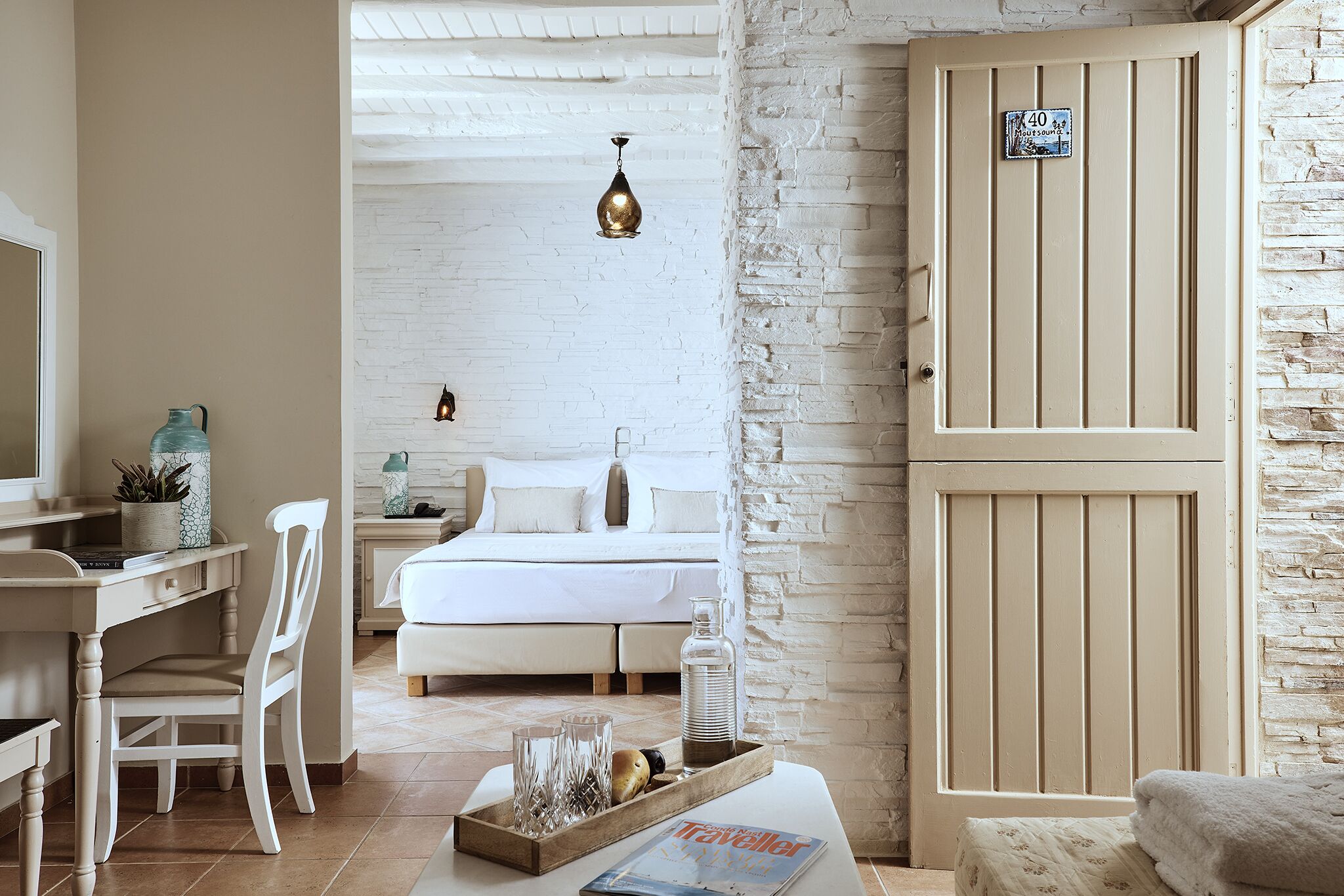 A hotel room with a bed, a desk with a chair, a bedside table with a lamp, and a door with a number sign.