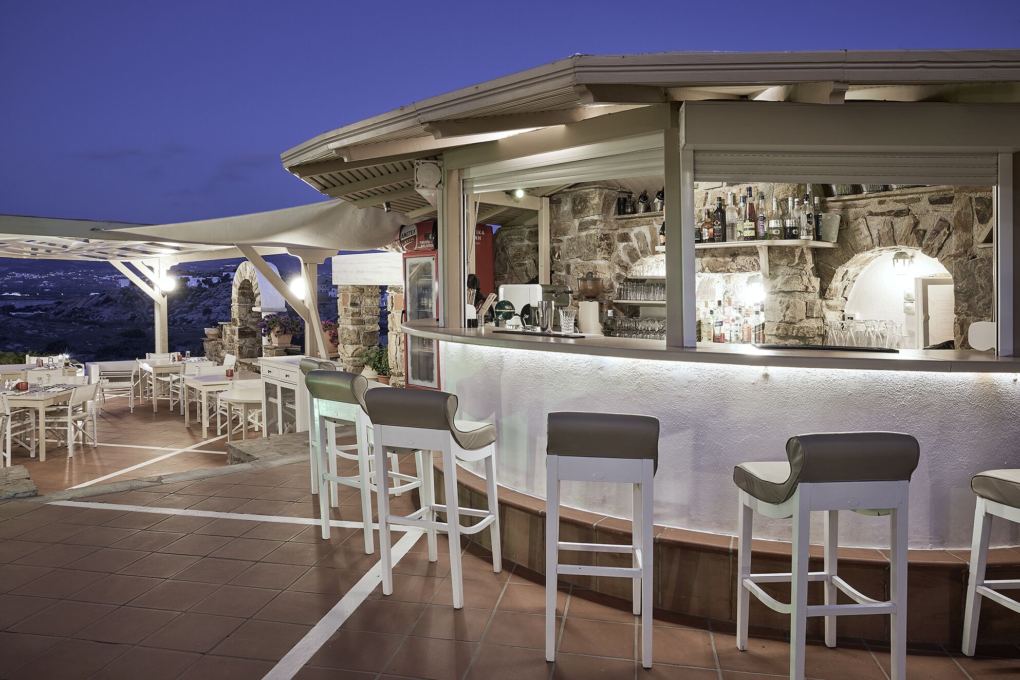 Zona de bar al aire libre con taburetes blancos y un muro de piedra de fondo.