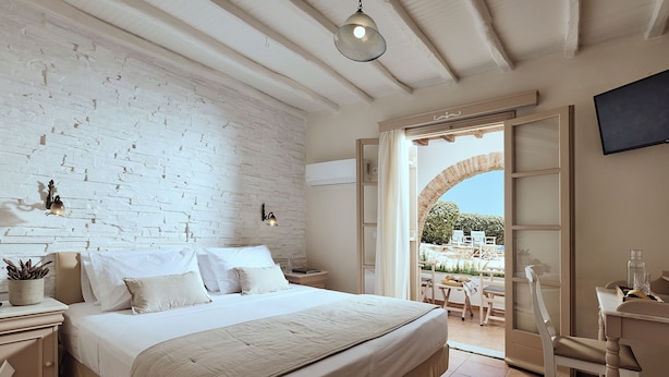 A bedroom with a large bed, a white brick wall, a ceiling fan, and a view of the outside patio. - Naxos Magic Village Hotel (Naxos)