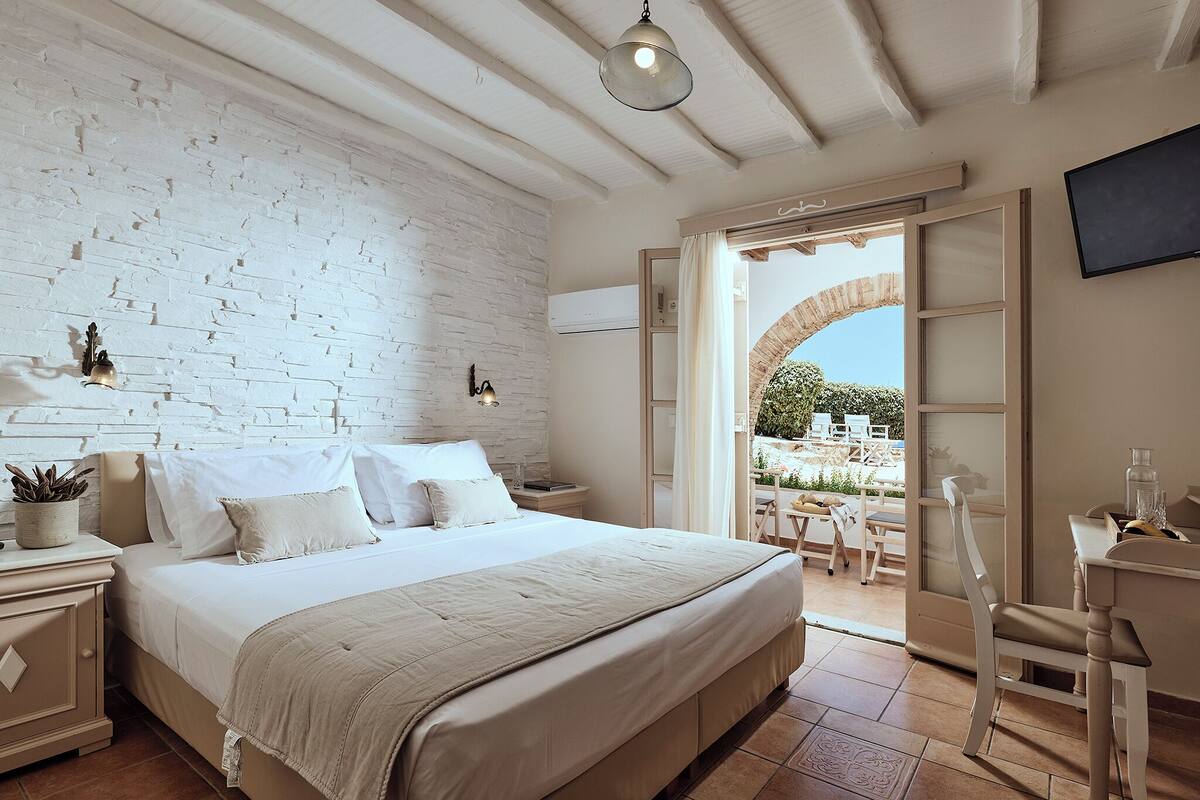 A bedroom with a large bed, a white brick wall, a ceiling fan, and a view of the outside patio.