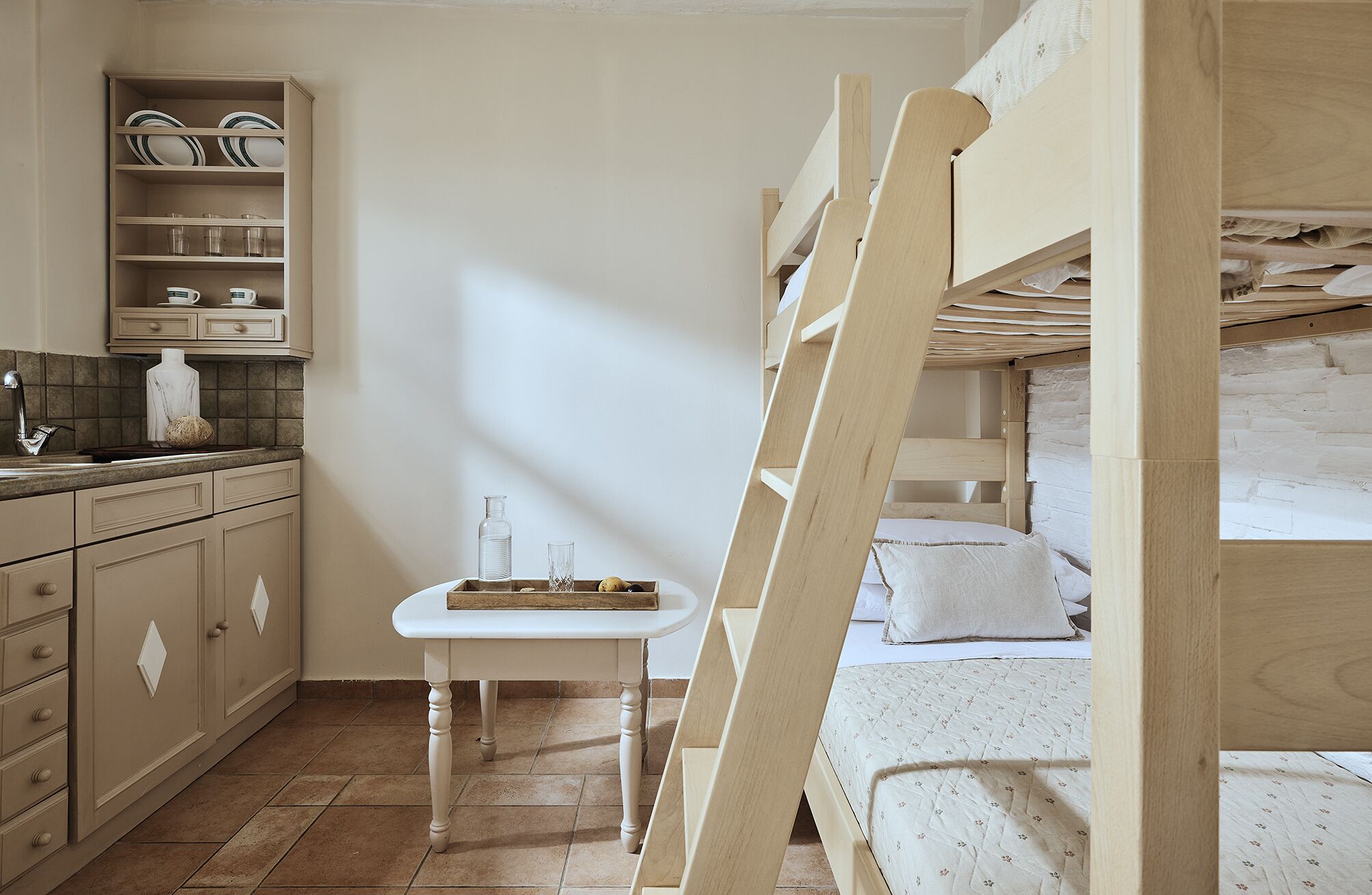 Una habitación con literas, una mesita con una silla, una zona de cocina con armarios y un lavabo, y suelo de baldosas.