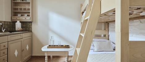 Una habitación con literas, una mesita con una silla, una zona de cocina con armarios y un lavabo, y suelo de baldosas.