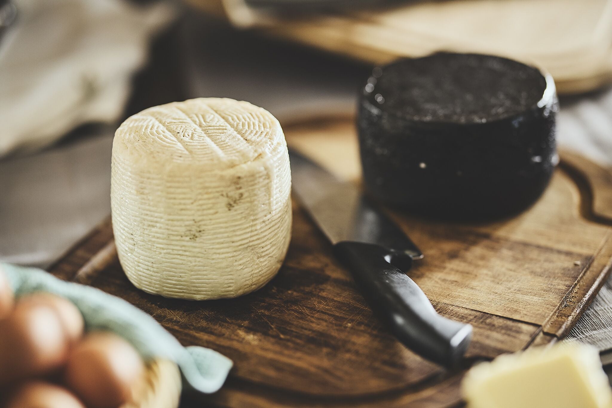 A cheese wheel, a black container, and a knife on a wooden board.