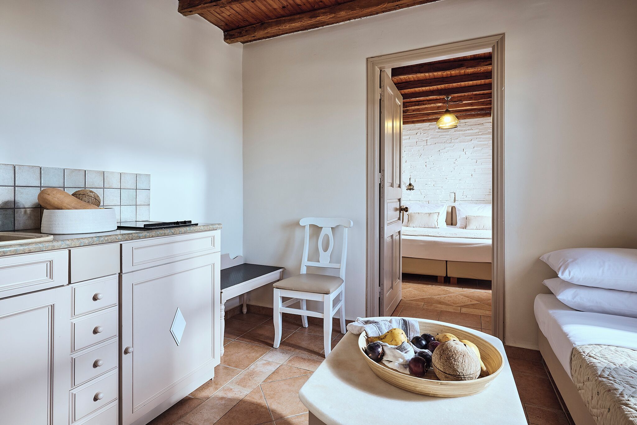 A room with a white bed, a wooden ceiling, a white chair, and a table with a fruit bowl.