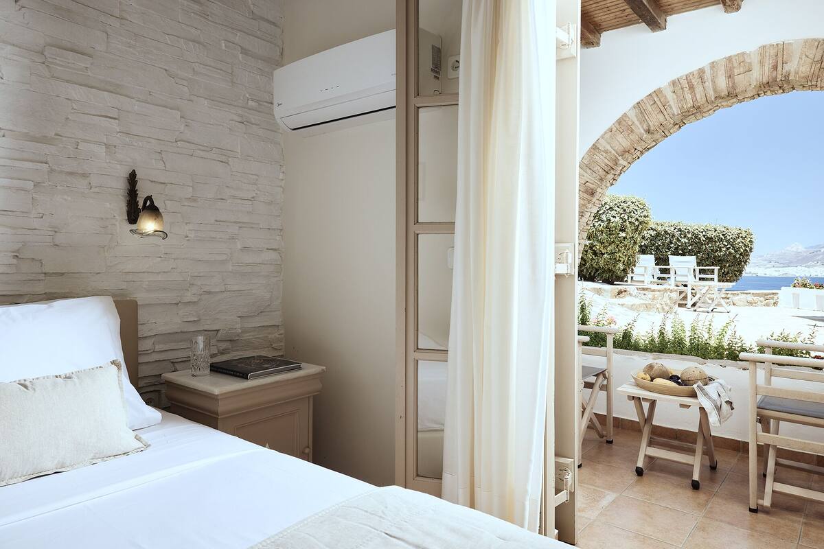 A bedroom with a white bed, a nightstand, and a view of a balcony with outdoor furniture.