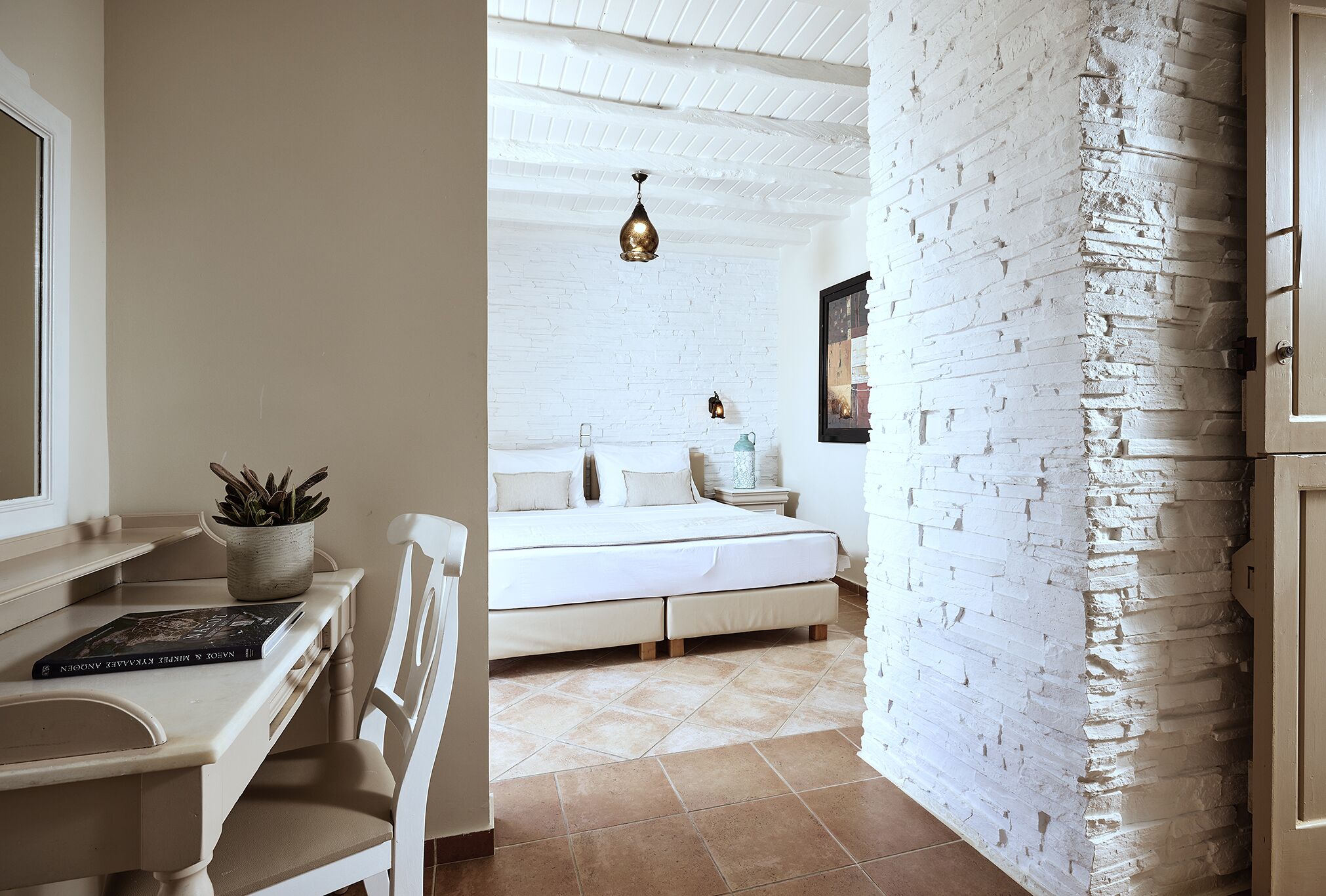 Ein Schlafzimmer mit weißer Ziegelwand, einem Bett mit weißer Bettwäsche, einem Nachttisch mit Lampe und einem Blumentopf auf dem Nachtkästchen.