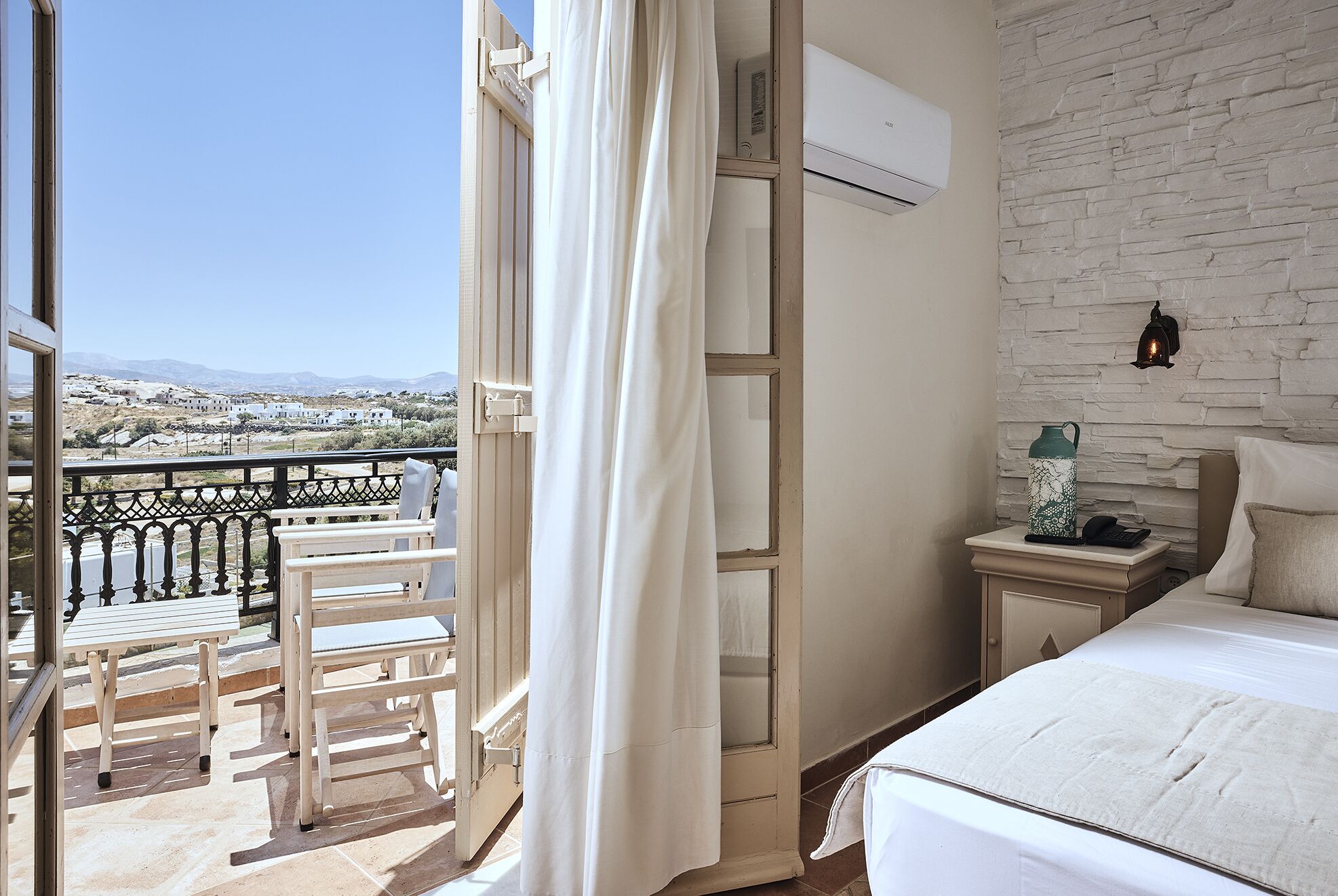 A balcony with a view of a hilly landscape, a bed with a white headboard, and a small bedside table with a lamp and a water bottle.