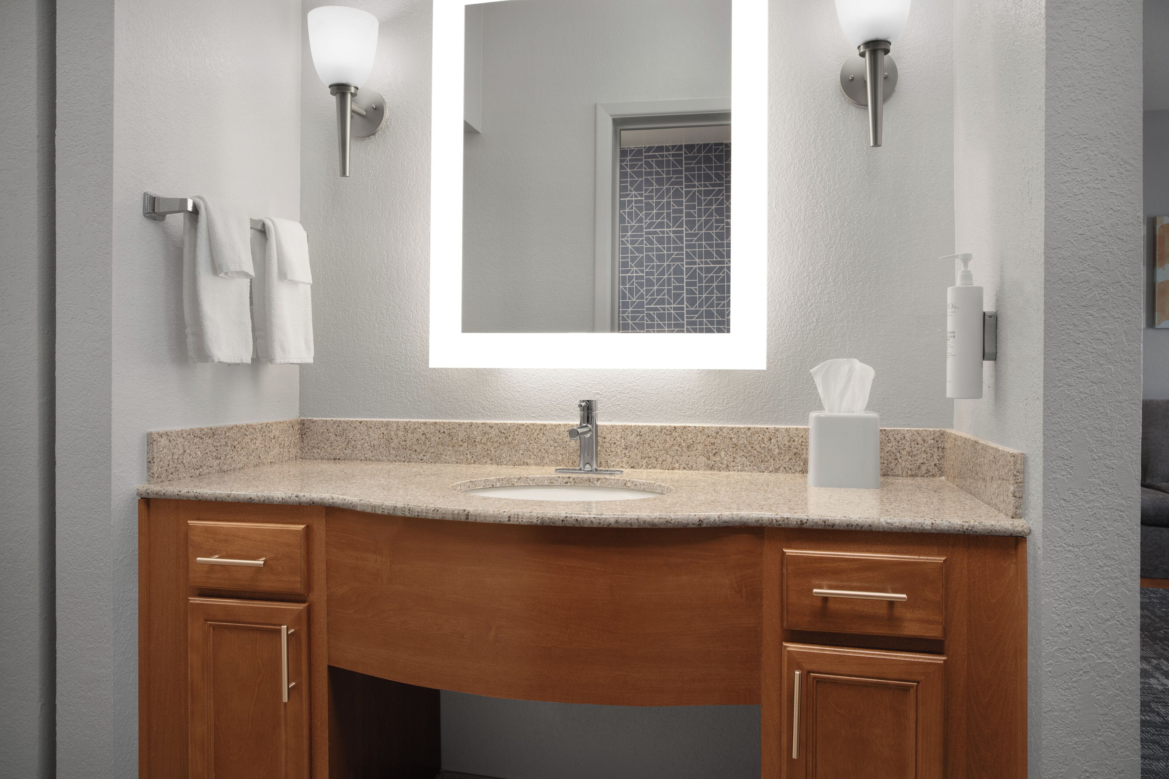 A bathroom with a granite countertop sink, wooden cabinets, a large mirror, and wall-mounted lighting.