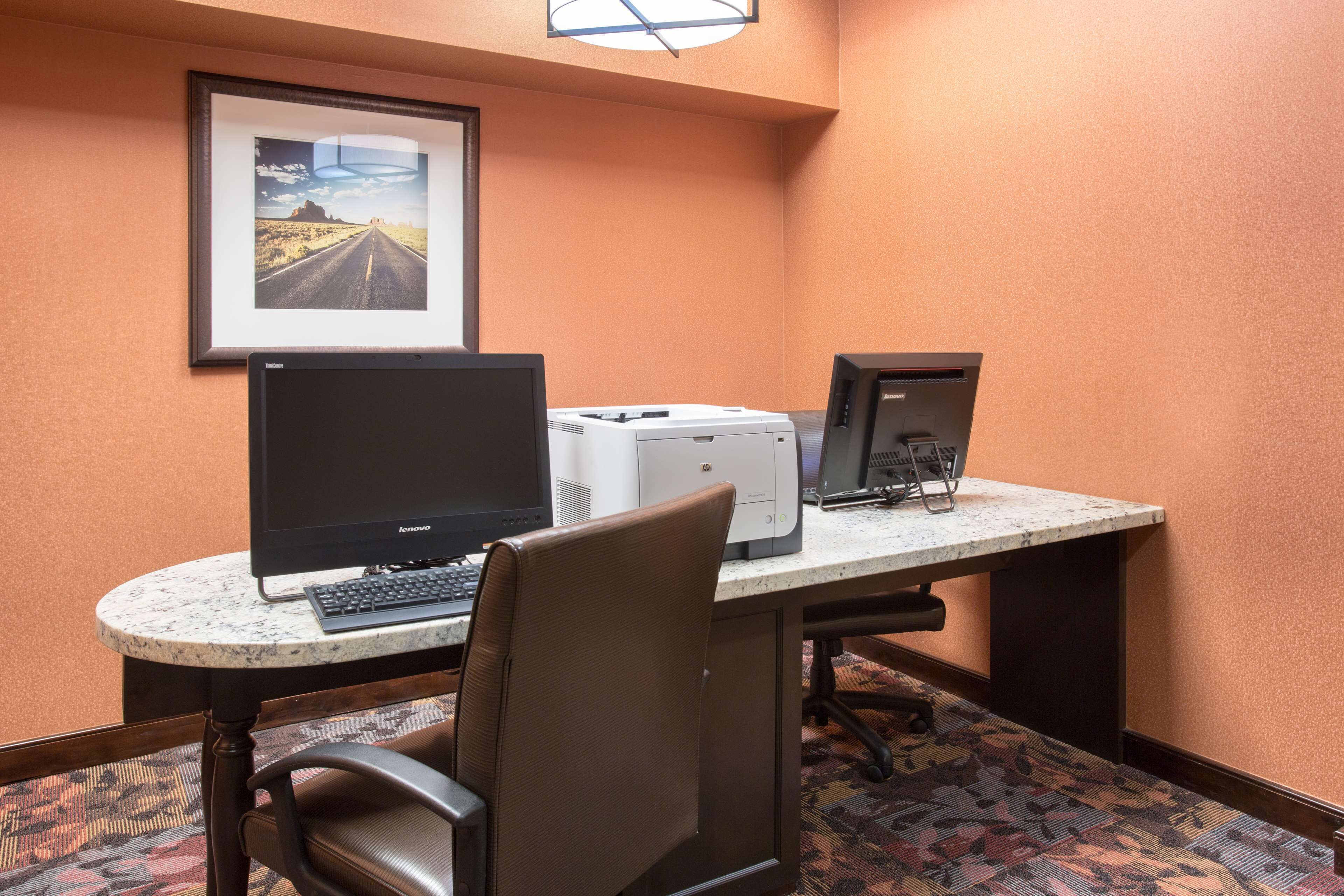 A hotel room with a desk, two monitors, a printer, and a framed picture on the wall.