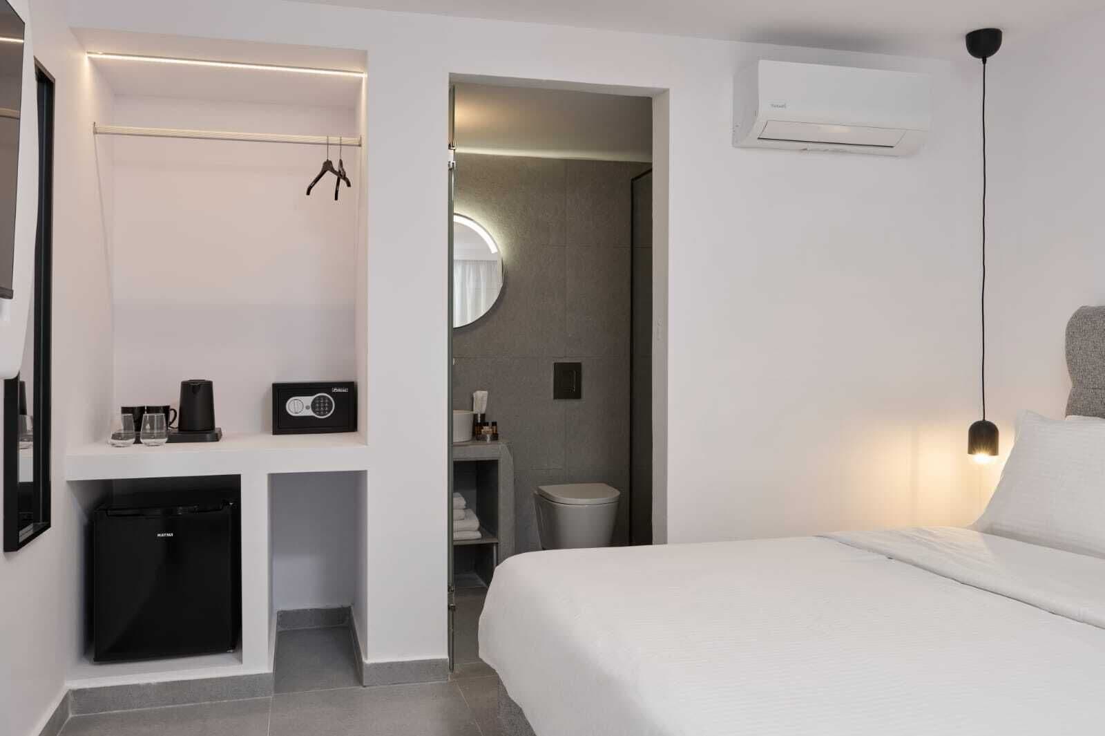 A modern hotel room with a bed, a small kitchenette with a coffee maker, a mirror, and an air conditioning unit.