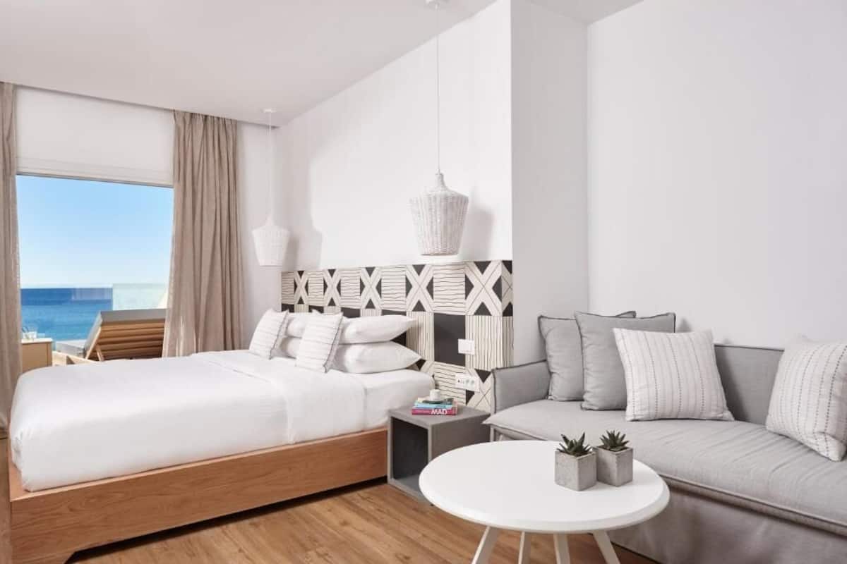 A modern hotel room with a bed, a sofa, a small table, and a view of the sea.