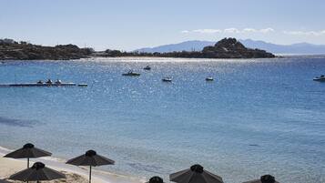 Une plage avec des parasols et des chaises longues, des bateaux sur l’eau et des montagnes en arrière-plan.
