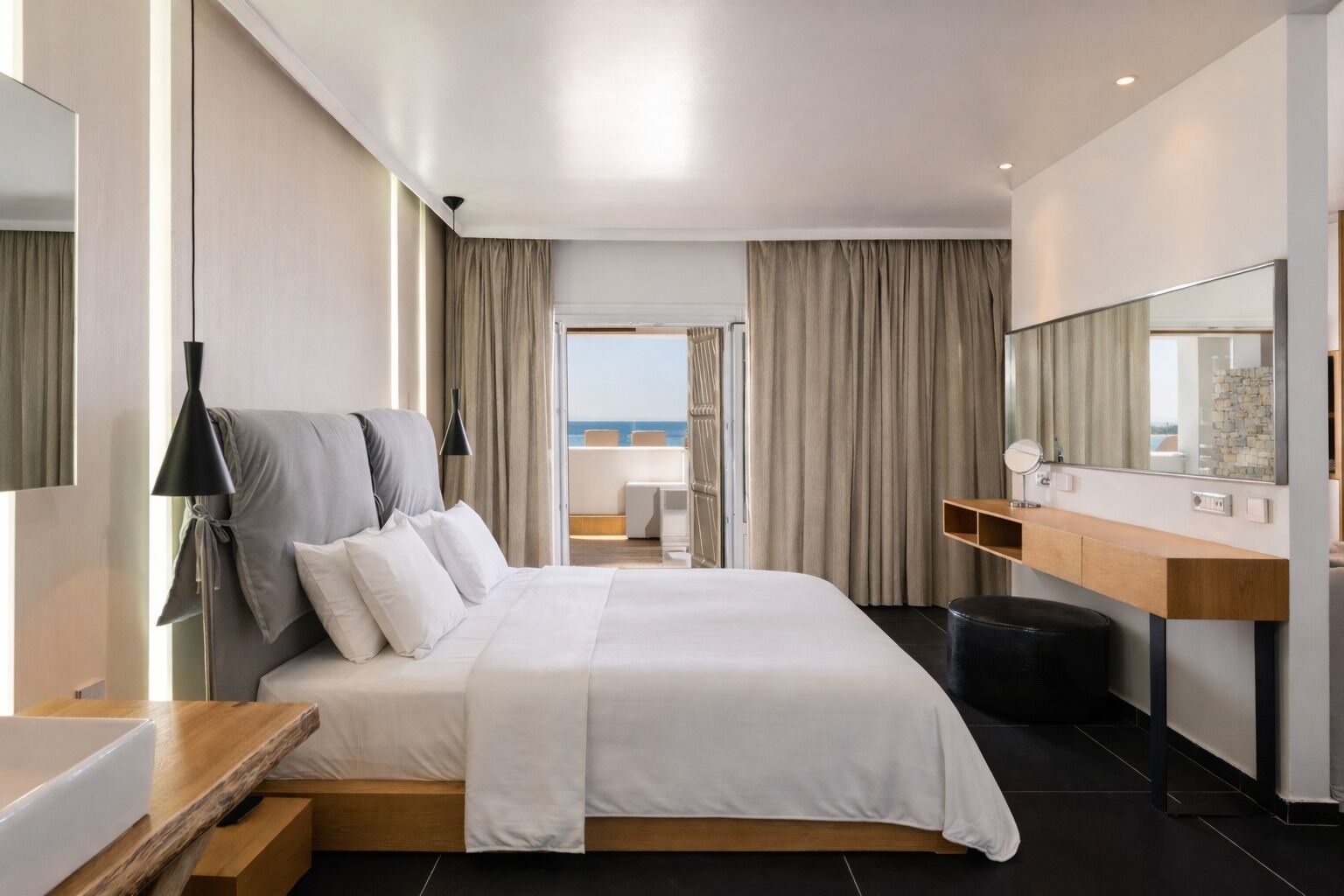 A modern hotel room with a bed, a wooden bedside table, a mirror, and a view of the sea.