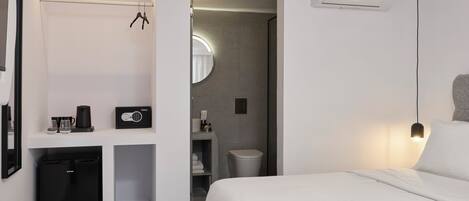 A modern hotel room with a bed, a small kitchenette, a mirror, and a built-in air conditioning unit.