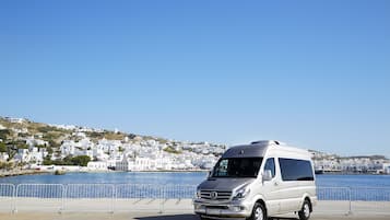 Una camioneta blanca estacionada frente a la costa, con edificios blancos al fondo y un cielo azul despejado.