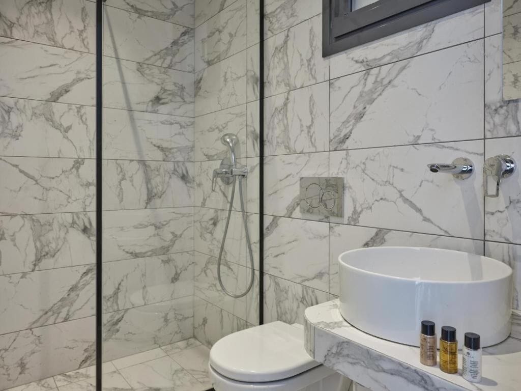 A bathroom with a marble-tiled shower, a toilet, and a sink with toiletries.