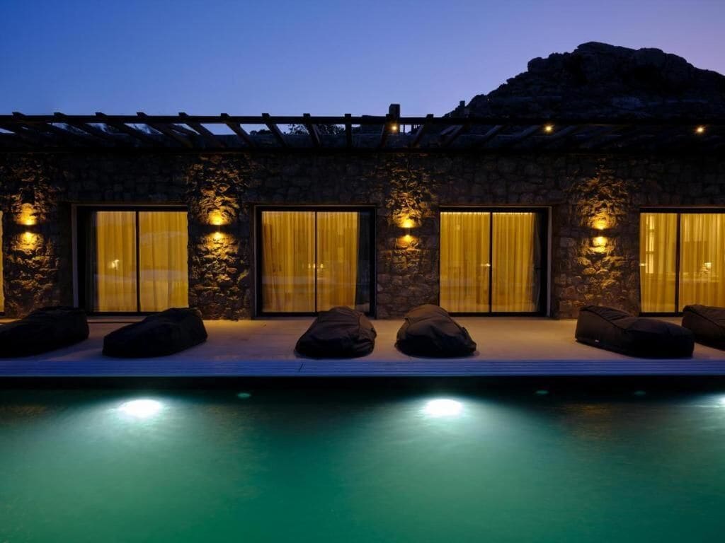 A modern building with stone walls, large windows with curtains, and a pool area with seating.