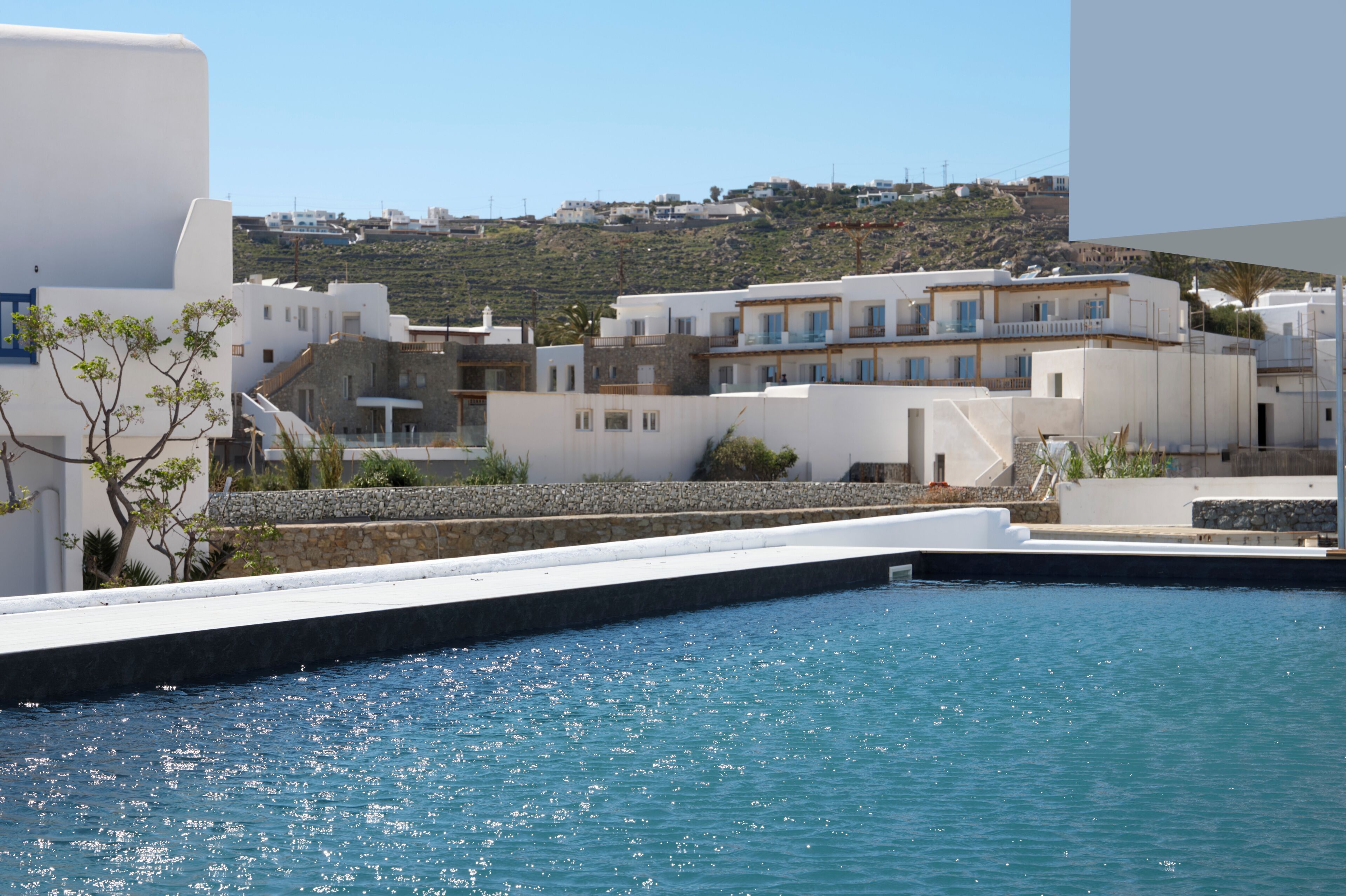 A coastal hotel with white buildings, a swimming pool, and a view of the hillside.
