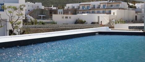 A coastal hotel with white buildings, a swimming pool, and a view of the hillside.