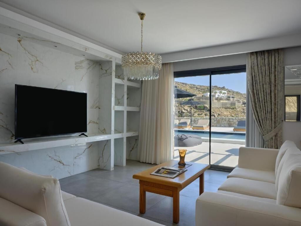 A modern living room with a flat-screen TV, a wooden coffee table, and a sofa. There is a large window with a view of a pool and hills.