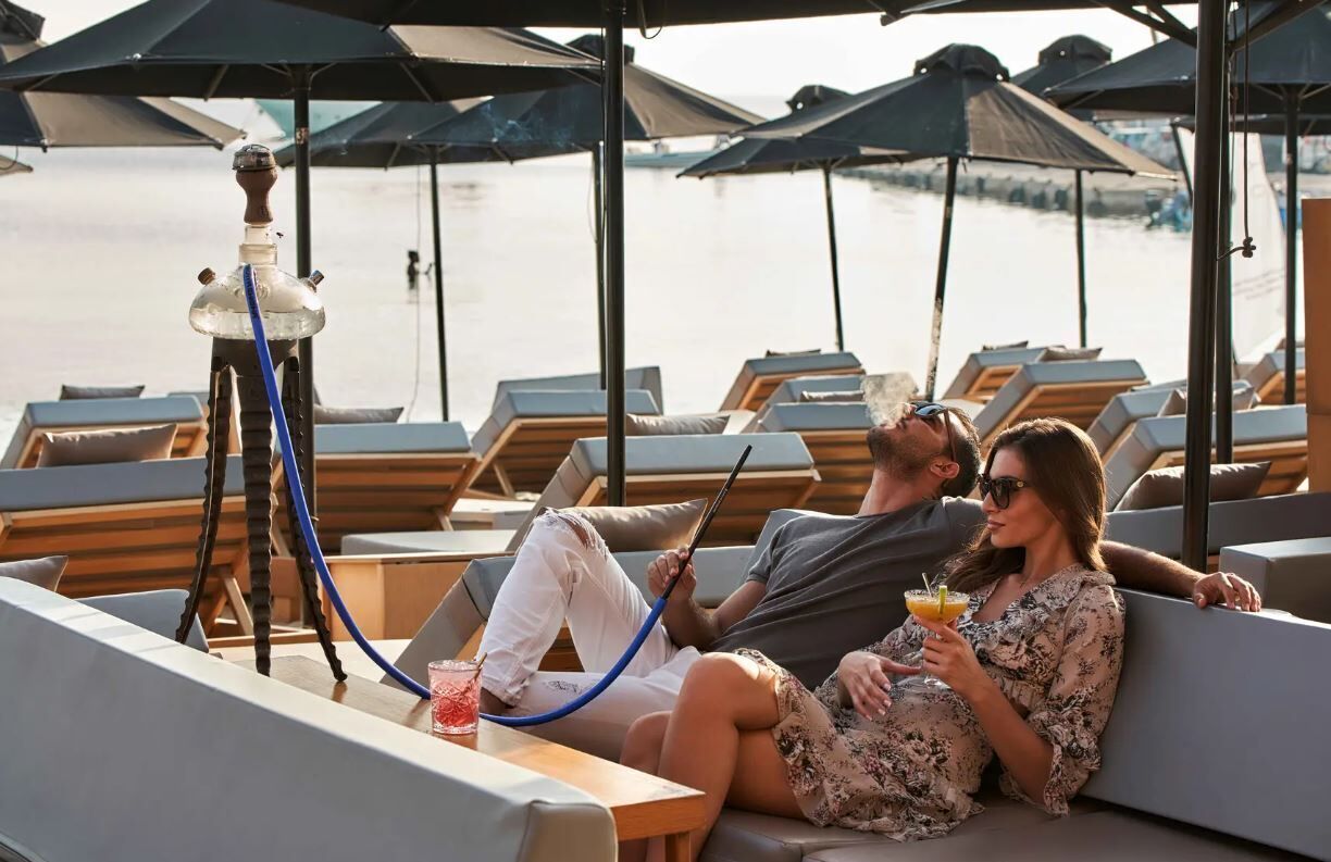 Ein Paar entspannt auf einer Liege am Wasser, mit einer Shisha und Getränken auf dem Tisch.