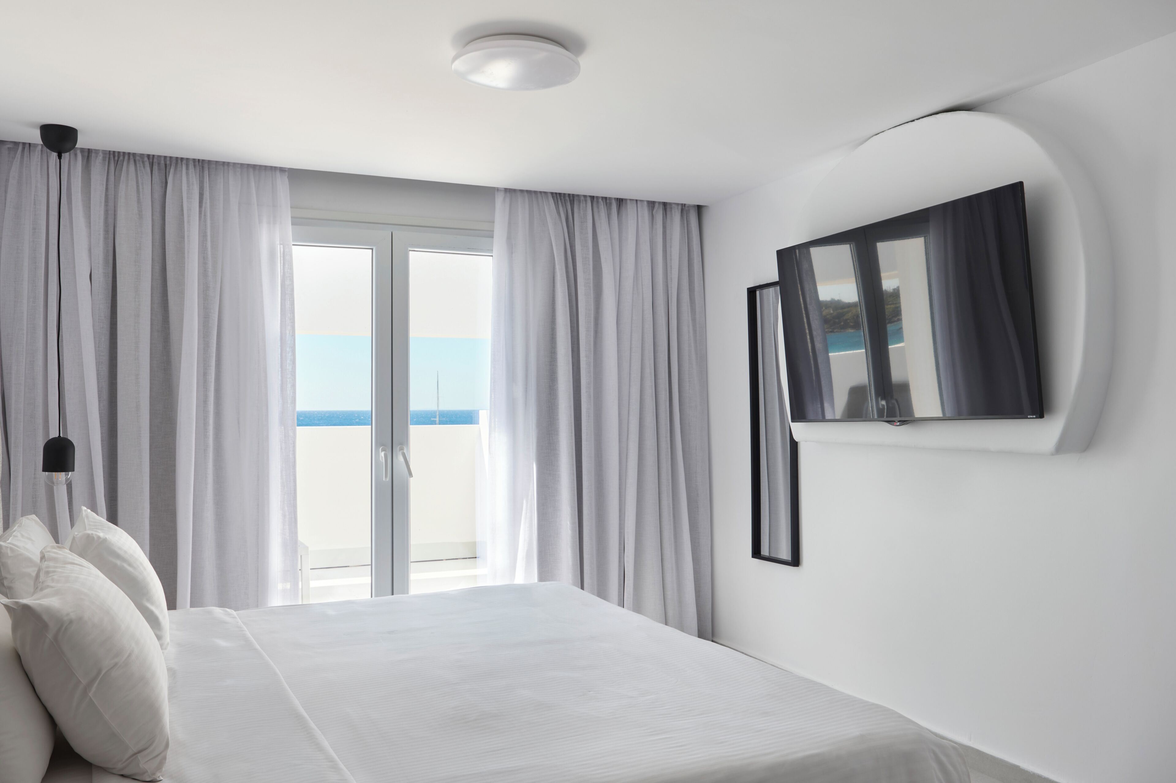 A hotel room with a bed, a large window with curtains, a ceiling light, and a wall-mounted TV.