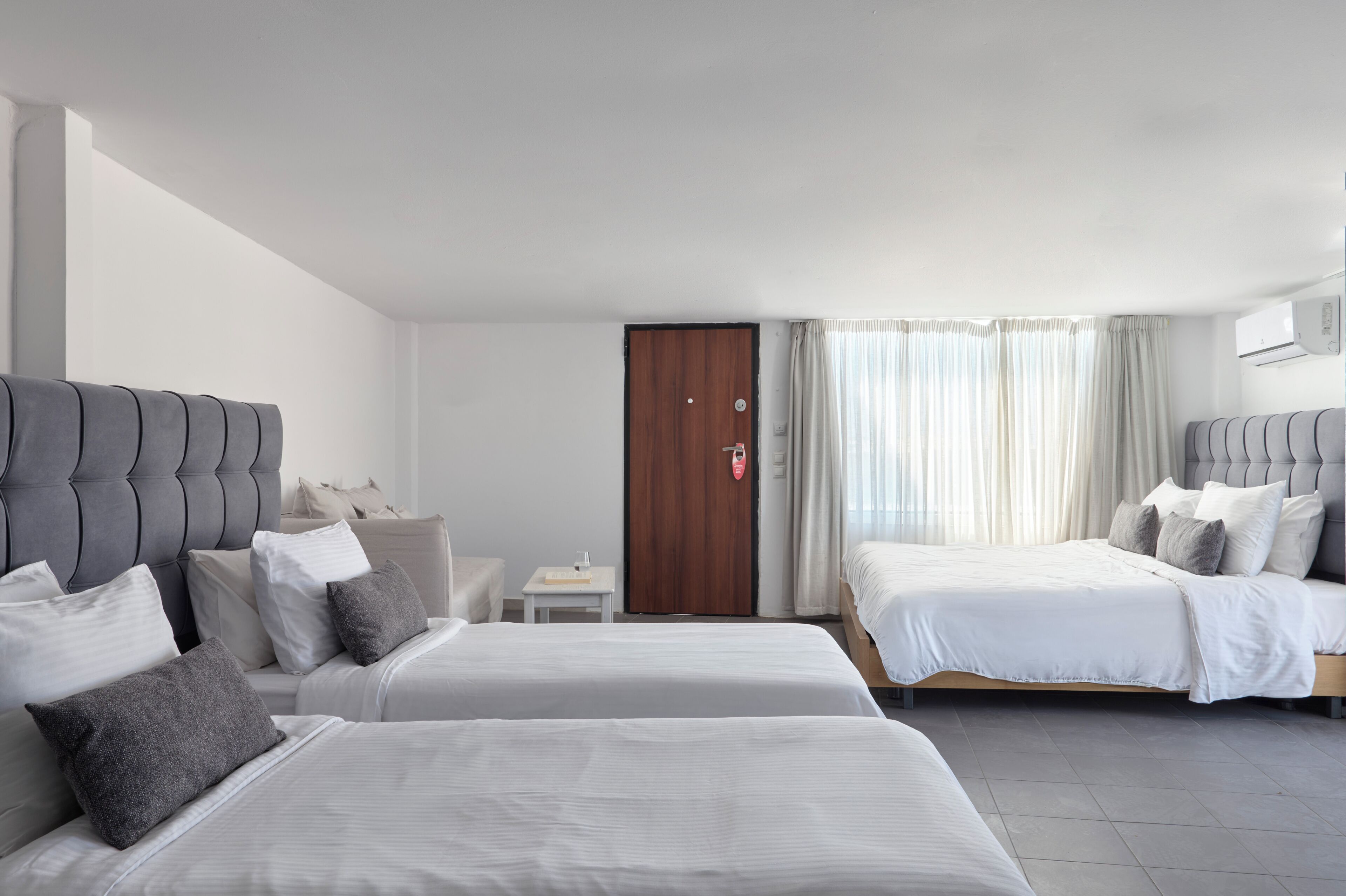 Habitación de hotel con dos camas, una mesita, una mesita de noche, una puerta de madera y una ventana con cortinas.