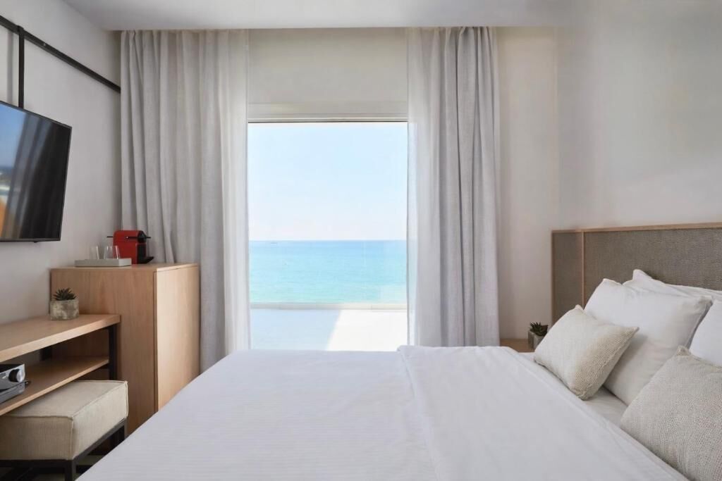 A hotel room with a bed, a desk, a TV, and a view of the sea.