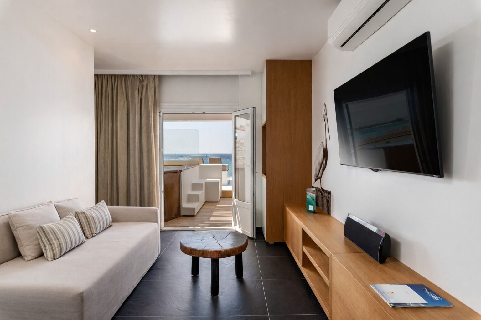 Een moderne hotelkamer met een bank, een klein rond tafeltje, een televisie en uitzicht op de oceaan door een open deur.