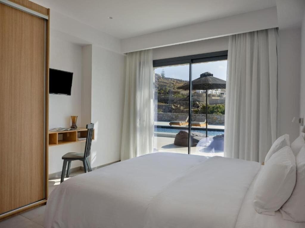 A hotel room with a bed, a TV, a desk with a chair, and a view of a pool and mountains.