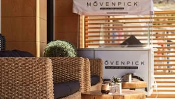 A wicker lounge chair with a cushion, a small round table with a drink and a plant, and a sign for 'MÖVENPICK' in the background.
