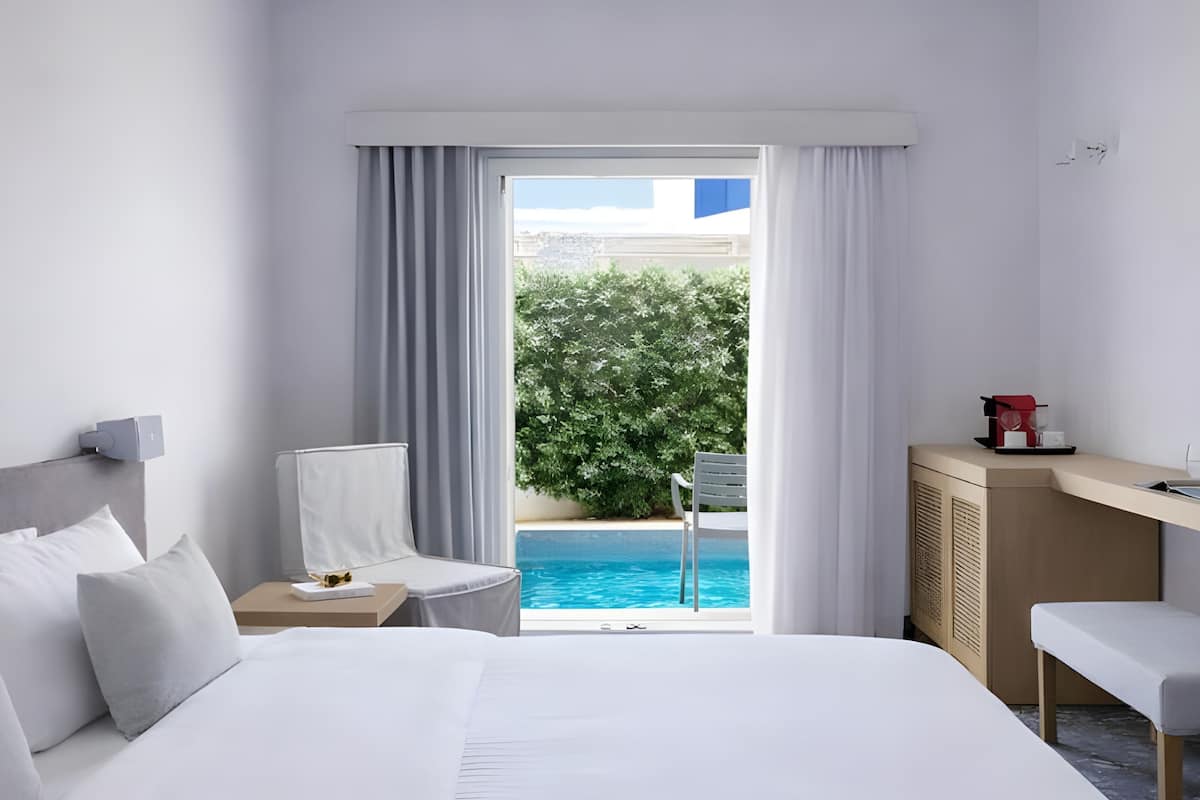 A hotel room with a bed, a desk, a chair, a coffee machine, and a view of a swimming pool and greenery.