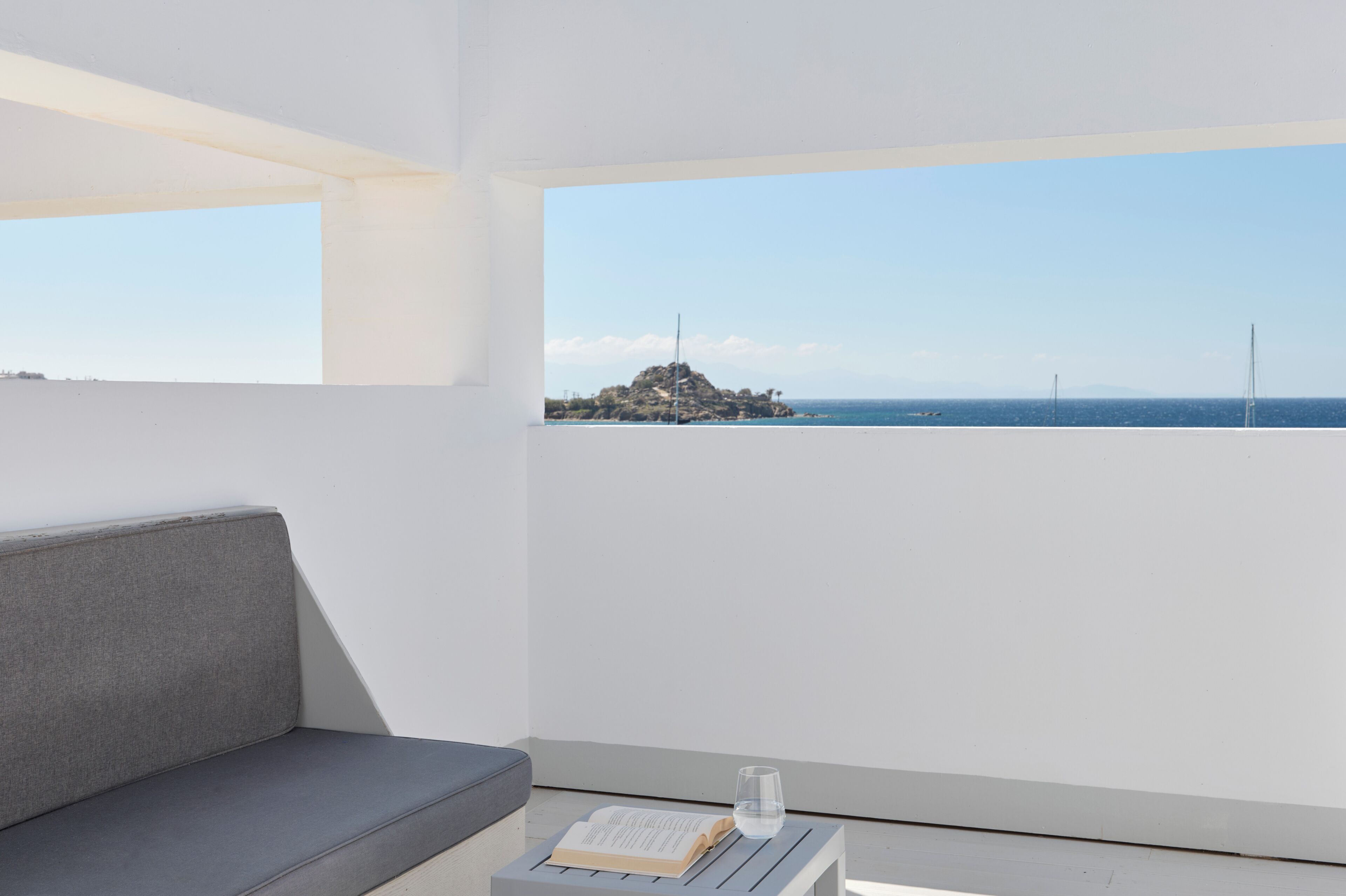 Una habitación moderna con un sofá gris, una mesita con un libro y un vaso, y un ventanal con vistas a un paisaje costero.