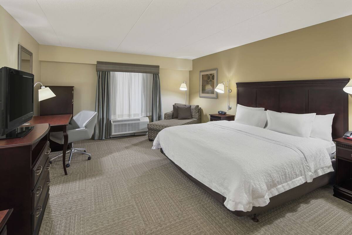 A hotel room with a large bed, a desk with a computer, a TV, a chair, and a window with curtains.