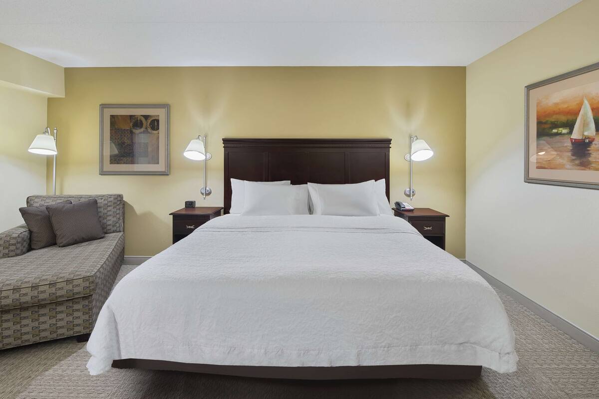 A neatly arranged hotel room with a large bed, two bedside tables with lamps, a sofa with cushions, and a framed painting on the wall.