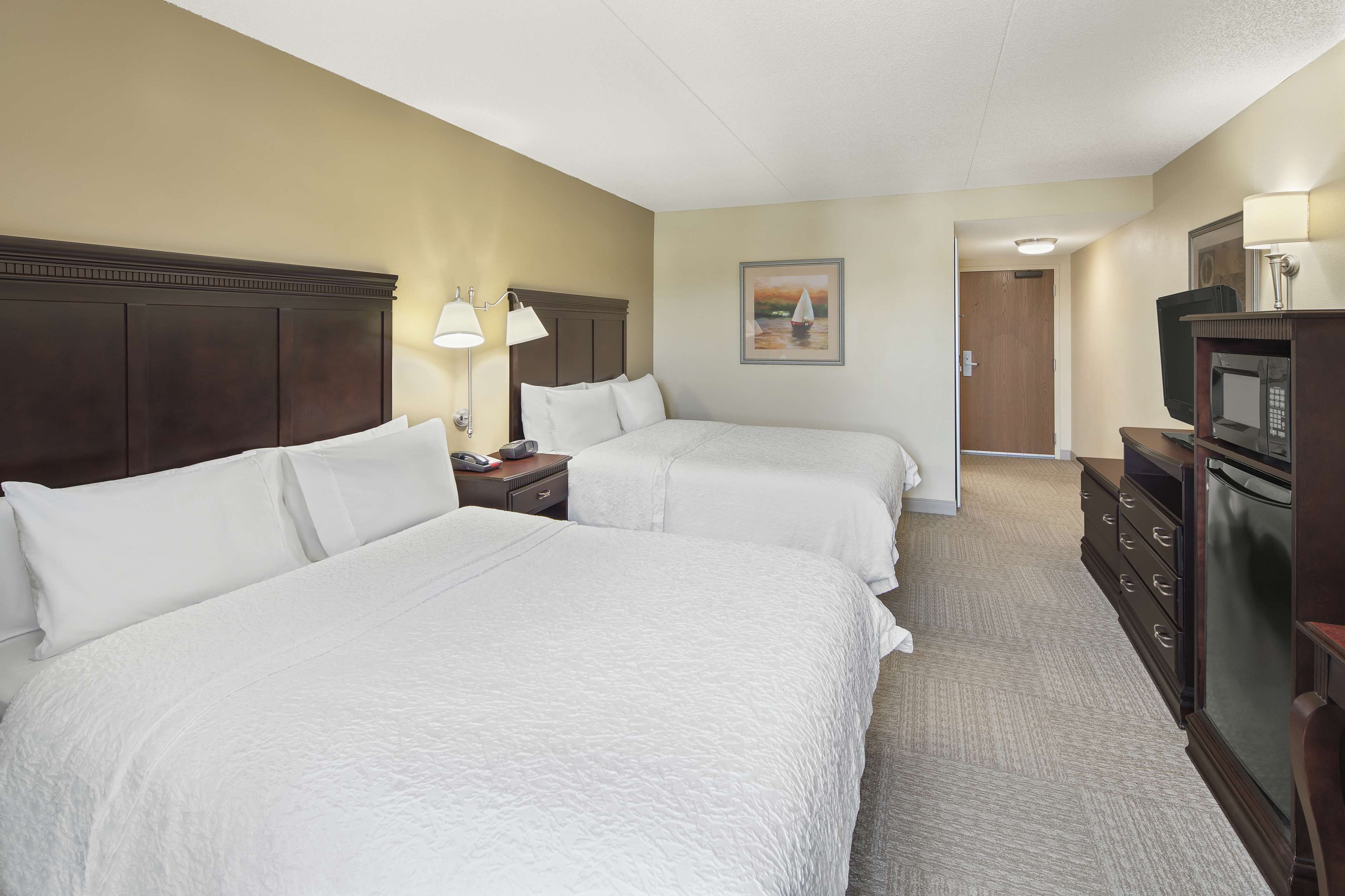 Quarto de hotel com duas camas, uma mesinha de cabeceira com luminária, cômoda, televisão e um quadro na parede.