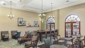 A hotel lobby with multiple seating areas, including round tables and chairs, a reception desk, and a large arched window offering a view outside.