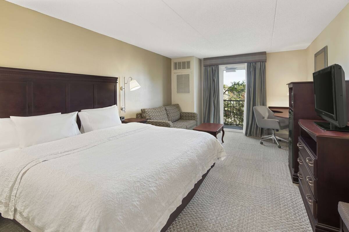 A hotel room with a large bed, a desk, a television, and a balcony with curtains.