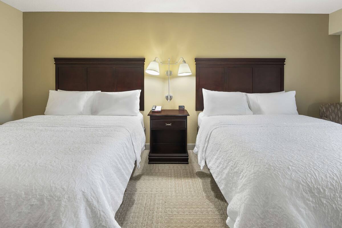 A hotel room with two beds, a nightstand with a lamp, and a wall-mounted light fixture.