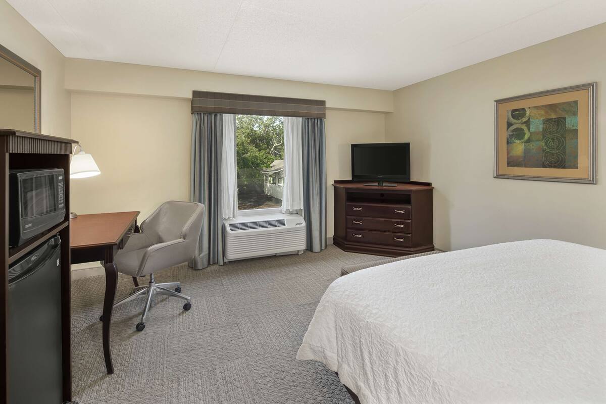 A hotel room with a bed, a desk with a chair, a television, a dresser, and a window with curtains.