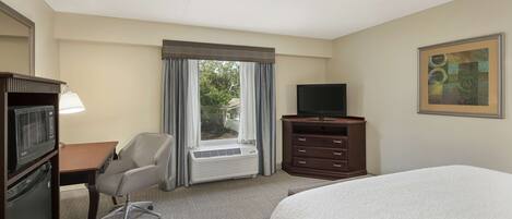 A hotel room with a bed, a desk with a chair, a television, a dresser, and a window with curtains.